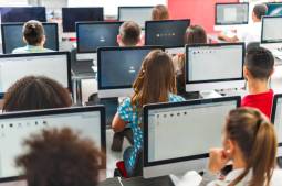 A photograph of a computer lab taken from behind several rows of students sitting in front of large computer monitors.